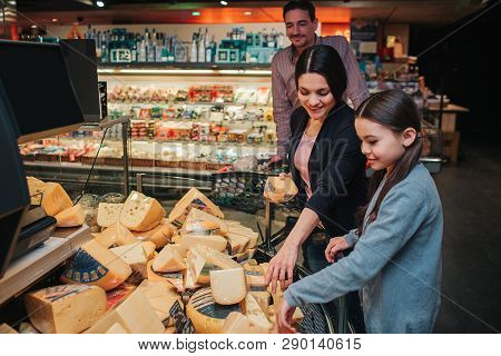 Young Parents And Daughter In Grocery Store. Mother Help Daughter To Pick Up Cheese. Father Stand Be