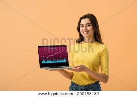 Cheerful Brunette Girl Holding Laptop With Traiding Website On Screen Isolated On Orange