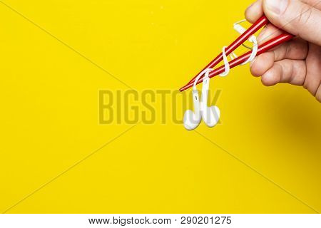 Hand  Is Holding A Headphones Using Chopsticks On A Bright Yellow Background, With Copy Space. Cconc