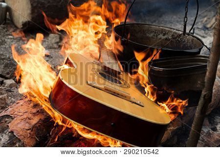 Guitar In Flame And Old Sooty Cauldrons On Campfire At Forest