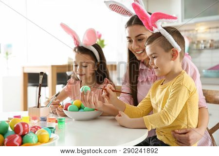 Mother And Her Children With Bunny Ears Headbands Painting Easter Eggs In Kitchen, Space For Text
