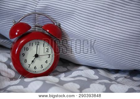 Retro Red Alarm Clock Stands On The Bed. Pillow In A Strip Of Gray And White.