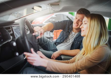 Young Family Buying First Electric Car In The Showroom. Attractive Smiling Couple Looking At Buttons