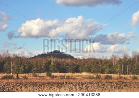 Autumn Or Spring Landscape With Dark Hill And Stones In Front, Trees And Blue Sky With Clouds In Gen