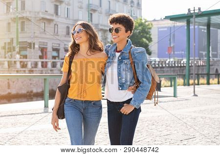 Two friends having a casual chat while walking on street. Two multiethnic girls laughing and walking in the city centre. Brazilian young woman and stylish latin girl wearing sunglasses.