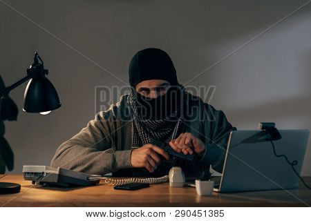 Terrorist In Black Mask Sitting At Table And Loading Gun