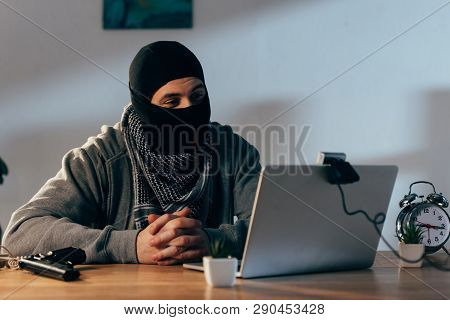 Terrorist In Mask Sitting With Interlaced Fingers And Looking At Webcam