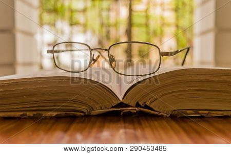 An Open Book And A Eyeglass Nicely Placed On Wooden Table On A Warm Sunny Morning, Side View Close U