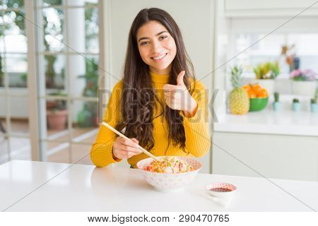 Young woman eating a bowl of Asian rice using chopsticks happy with big smile doing ok sign, thumb up with fingers, excellent sign