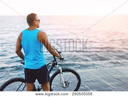 Back View Of Athletic Sexy Man With Bicycle Outdoors At Seaside. And Watching The Sunset