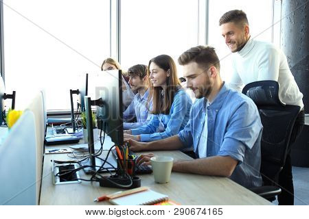 Group Of Young People In Casual Wear Sitting At The Office Desk And Discussing Something While Looki