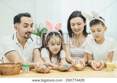 Inspired Family In White T-shirts Sitting Together At The Table, Holding Colored Easter Eggs In Thei