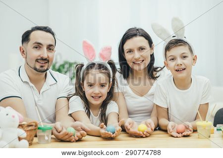 Inspired Family In White T-shirts Sitting Together At The Table, Holding Colored Easter Eggs In Thei