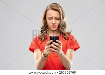 technology, cyberbullying and emotions concept - sad teenage girl in red t-shirt using smartphone over grey background