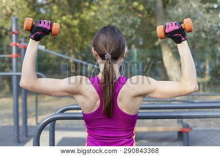 The Back View Of Slim Girl In Bright Sportswear Training With Dumbbells For Biceps At Outdoor Sportg