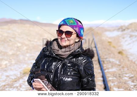 Smiling White Girl Wearing Sunglass, Black Coat, Brown Scarf, Colorful Band, And Holding A Pink Smar