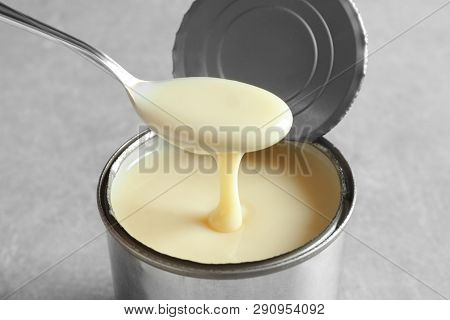 Condensed Milk Pouring From Spoon Into Tin Can On Grey Background, Closeup. Dairy Product