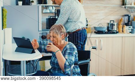 Disabled Senior Woman In Wheelchair Using Tablet Computer In Kitchen. Paralysied Handicapped Old Eld