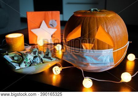 Carved Halloween Pumpkin With Sad Eyes Wearing Mask On The Table With Sweets, No Party During Covid 