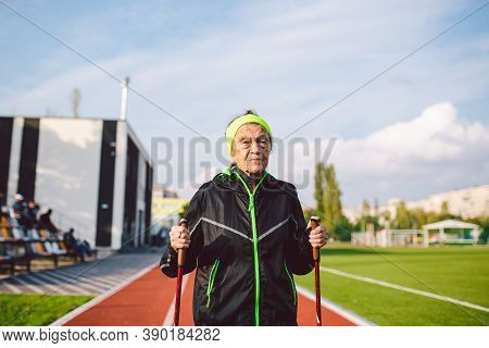 Senior Woman Walking With Walking Poles In Stadium On A Red Rubber Cover. Elderly Woman 88 Years Old
