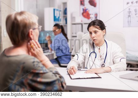 Young Doctor Examining Senior Patient In Hospital Cabinet Wearing White Coat And Stethoscope. Mature
