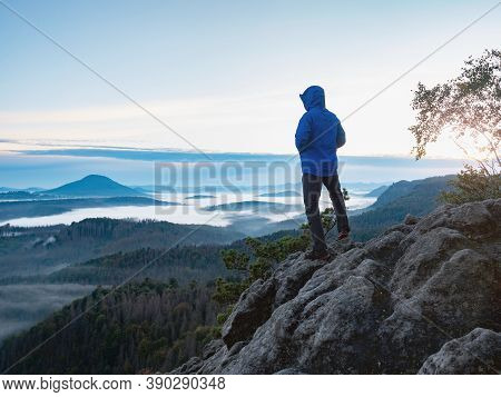 Hiking In Sunrise. Man Hiker Wear Hood And Blue Windcheater Stay Alone In Sunset At Mountain Edge. H