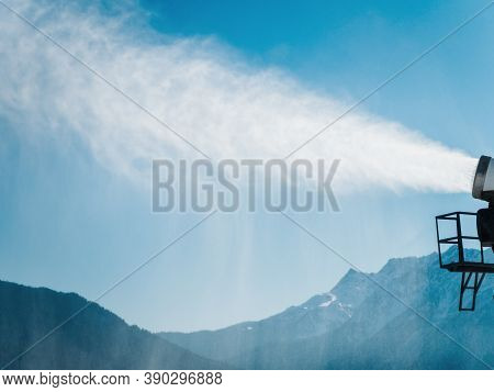 Artificial Snow Scatters From The Muzzle Of A Snow Cannon Against The Backdrop Of Blue Sky And Mount