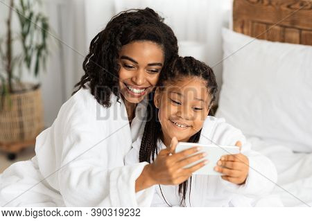Cute Little Black Girl And Her Mom Wearing Bathrobes And Taking Selfie On Smartphone Together, Prett