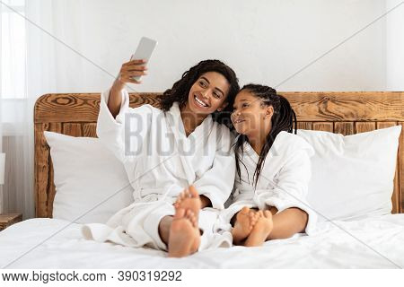 Spa Day Fun. Cheerful Black Mother And Little Daughter In Bathrobes Taking Selfie On Bed At Home, Re