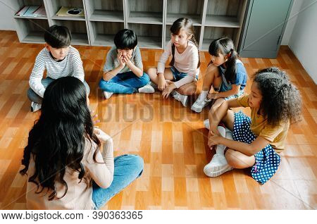 Elementary International Students Group Sit Cross Legged In Circle Around The Teacher And Listening 