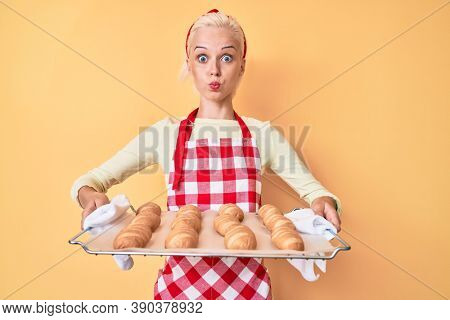 Young blonde woman wearing baker uniform holding homemade bread puffing cheeks with funny face. mouth inflated with air, catching air. 