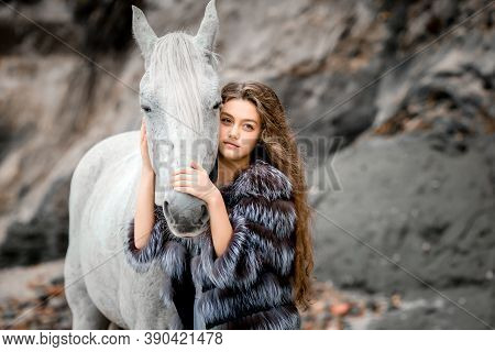 A Beautiful Young Girl Embraces A White Horse. Cool Weather, Teen Girl With Long Curly Hair In A Fur
