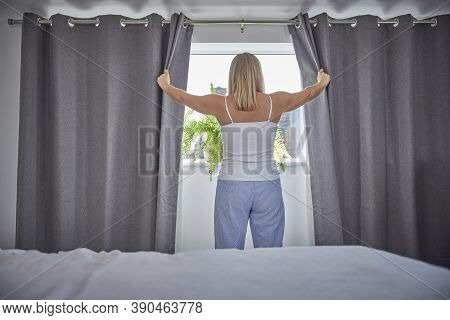 Rear View Of Woman Wearing Pyjamas Opening Curtains And Looking Out Of Window In Morning