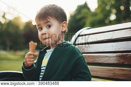Cute Boy Is Eating Ice Cream And Looking Surprised At Camera While Sitting On The Bench In Park