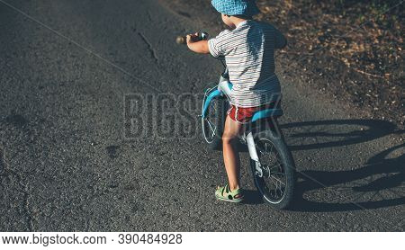 Caucasian Boy With Hat Is Learning To Ride The Bike On The Road During A Sunny Summer Day