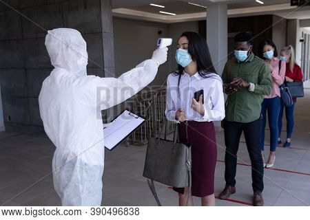 Multi ethnic group of colleagues having temperature taken by worker in protective suit before being allowed in to work. Hygiene and social distancing in workplace during Coronavirus Covid 19 pandemic.