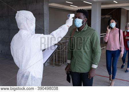 Multi ethnic group of colleagues having temperature taken by worker in protective suit before being allowed in to work. Hygiene and social distancing in workplace during Coronavirus Covid 19 pandemic.