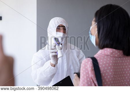 Mixed race businesswoman is shown her temperature by worker in protective suit before being allowed in to office. Hygiene and social distancing in workplace during Coronavirus Covid 19 pandemic.