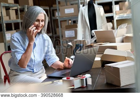 Older Female Fashion Dropshipping Business Owner Using Laptop Talking On Phone In Warehouse. Mature 