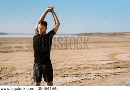 Motivated focused young sportsman exercising at the beach, stetching