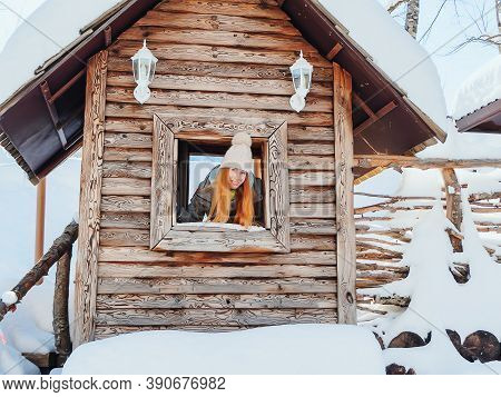 A Redhaired Girl Looks Out Of The Window Of A Decorative Wooden Hut On A Winter Day