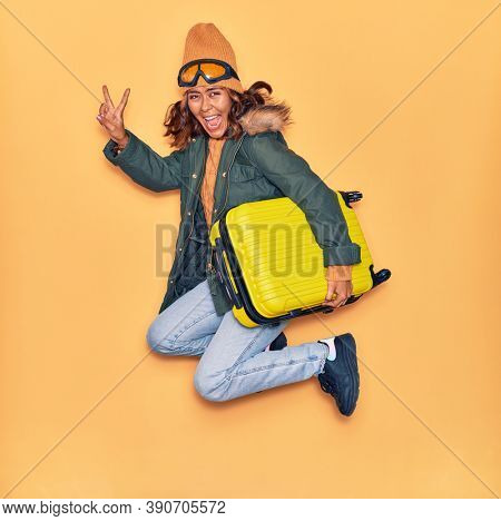 Young beautiful latin woman wearing winter clothes smiling happy. Jumping with smile on face holding cabin bag doing victory sign over isolated yellow background