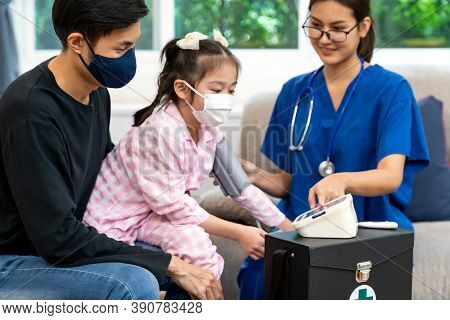 Asian female doctor take blood pressure measurement to little girl child sit with her father in living room while doctor visit at home. Home health care delivery and doctor visiting concept.