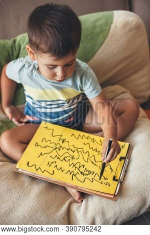 Cute Caucasian Boy Is Drawing Something On A Yellow Paper With A Marker While Sitting At Home In Bed