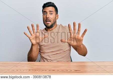 Handsome hispanic man wearing casual clothes sitting on the table afraid and terrified with fear expression stop gesture with hands, shouting in shock. panic concept. 