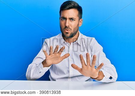 Handsome hispanic man wearing business clothes sitting on the table moving away hands palms showing refusal and denial with afraid and disgusting expression. stop and forbidden. 