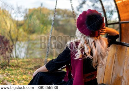A Woman Sits With Her Back On A Garden Swing, Tossed In A Burgundy Coat And In A Biret, With Beautif