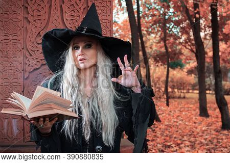 A Witch In A Hat Reads A Spell From A Book. Halloween, A Young Woman At The Carnival In The Guise Of