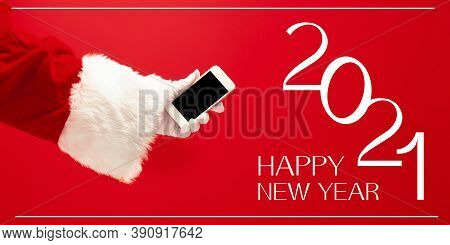 Hand Of Santa Claus Holding Phone, Greeting With New Year 2021 And Christmas. Red Background With Le