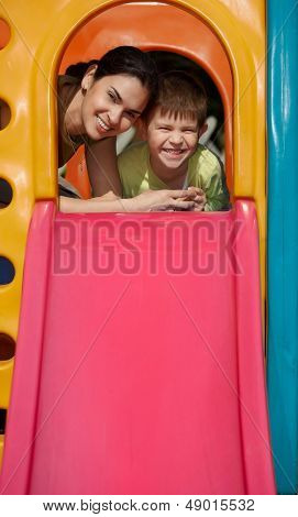 Mother and son at top of slide at playground, smiling, looking at camera.